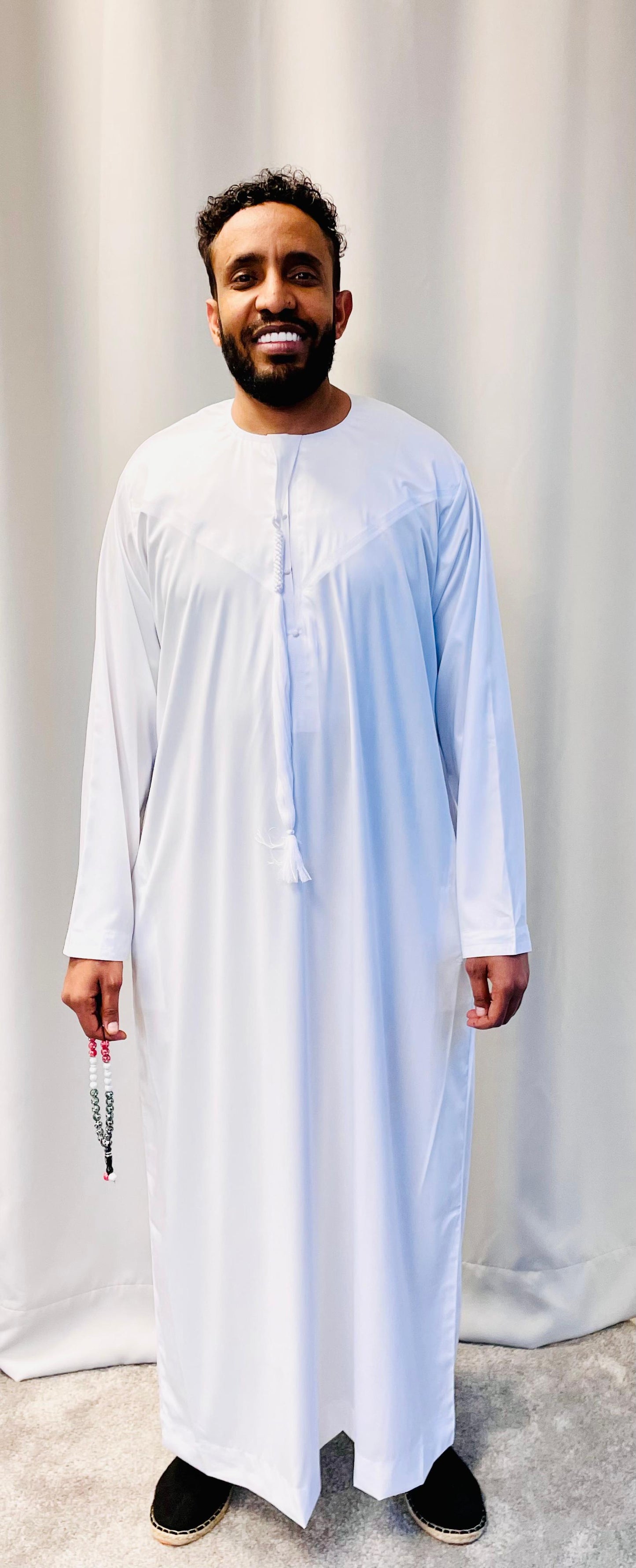 A man smiles in front of a light curtain, wearing the Shop Stylish Emirati Khamis Size 60 from Bihi Boutique and holding a beaded tasbih in his left hand.