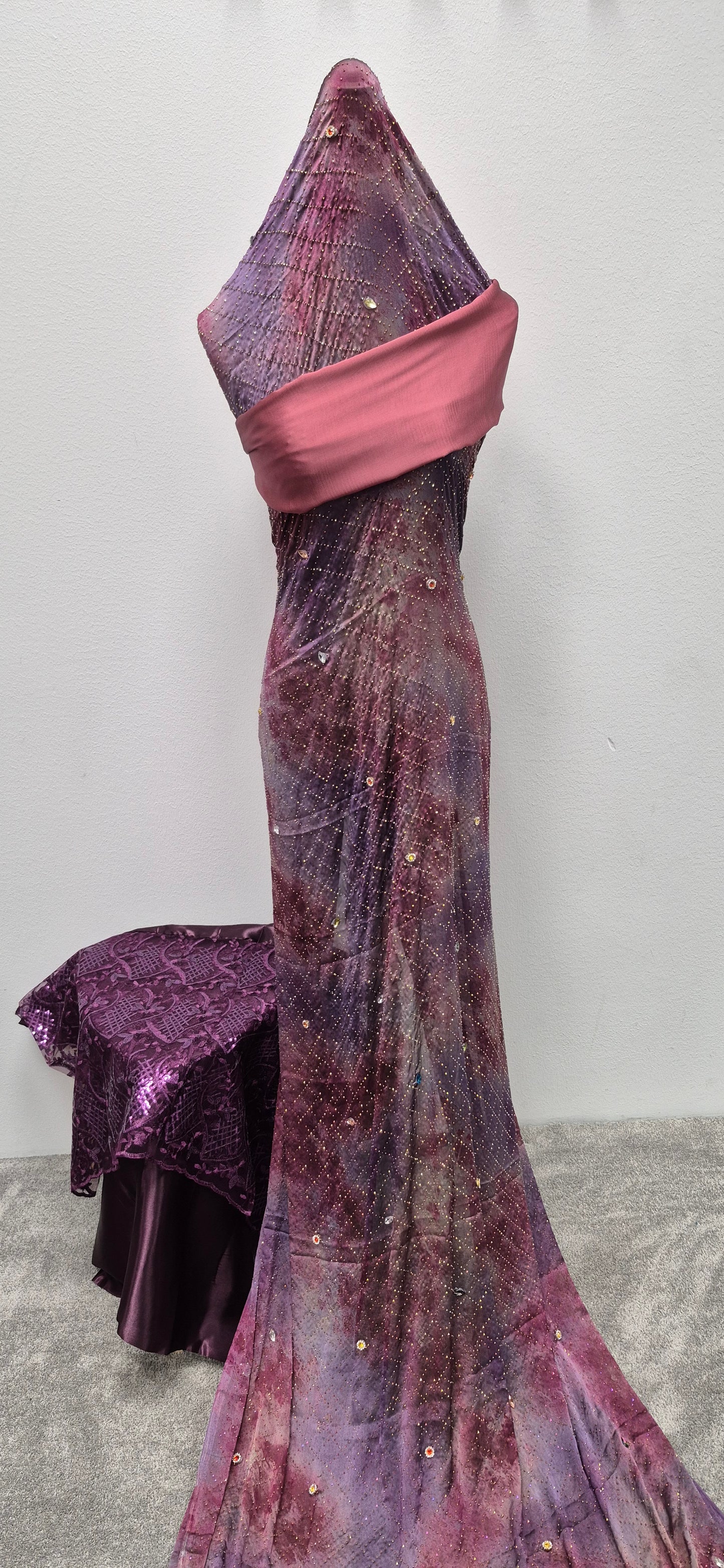 A mannequin showcases the Shop Stylish Fransawi Dirac Half Bac by Bihi Boutique—a shimmering purple and pink gown with sequins and a wide sash, fabric draped over the head. A matching purple cloth from Bihi Boutique covers a nearby table.