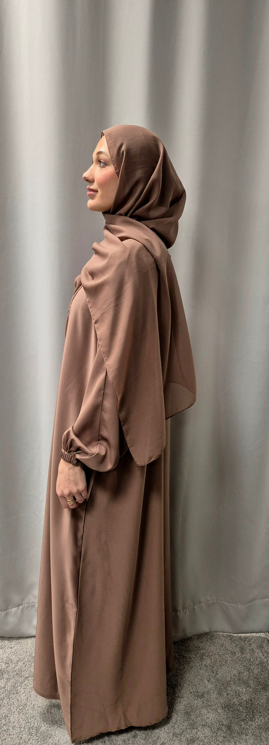 A woman models the Bihi Boutique Amina Rose Gold Abaya with matching chiffon hijab, size 58, standing sideways against a light gray curtain and gazing gently upward.