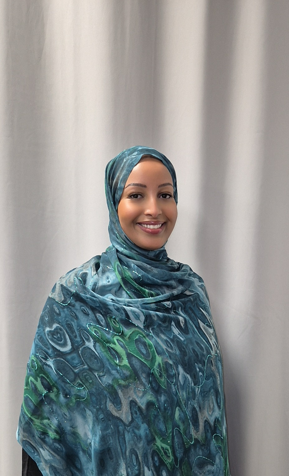 A woman wearing the Elegant Marble Chiffon Hijab from Bihi Boutique smiles while standing in front of a plain, light-colored curtain.