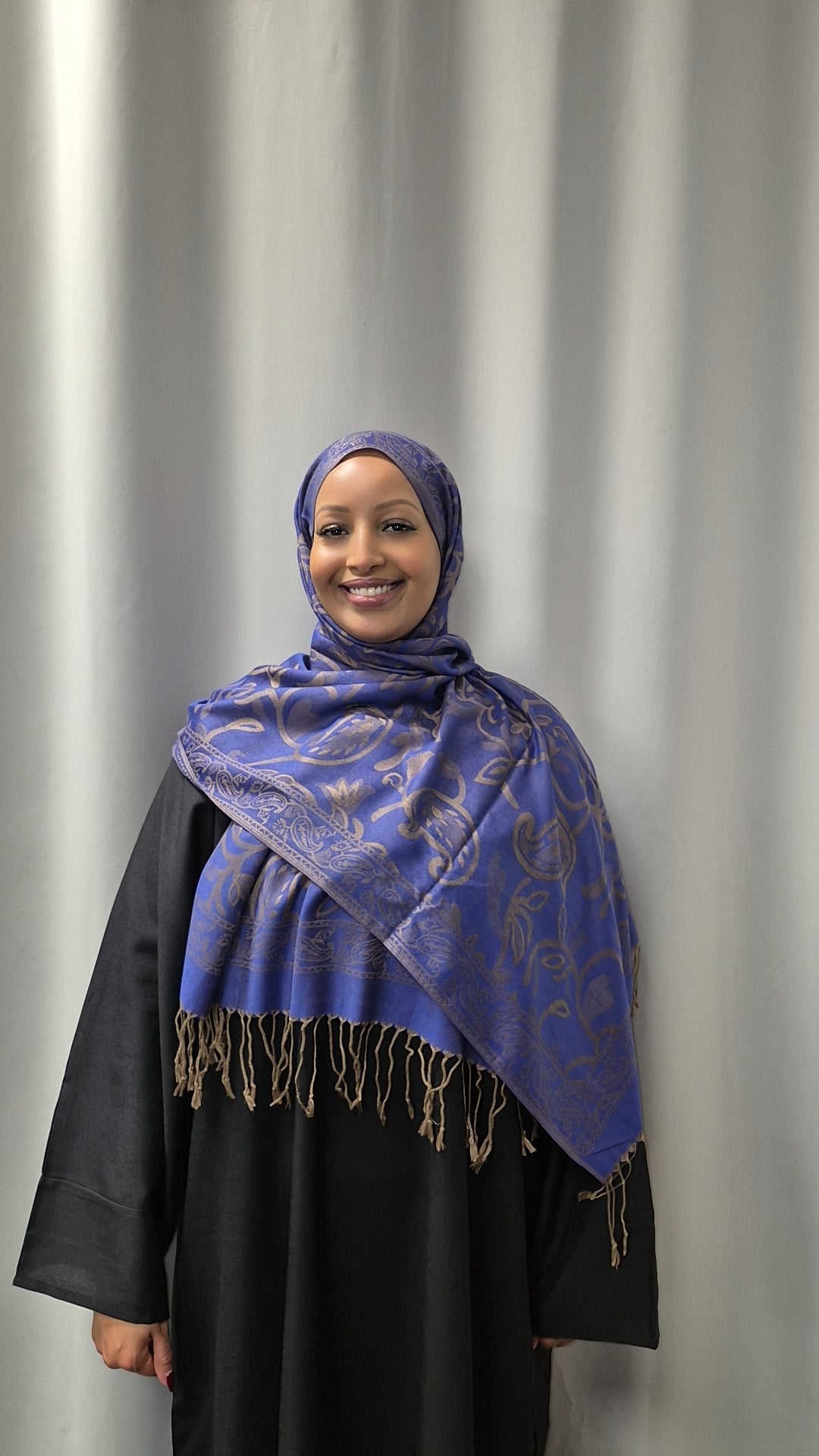 A woman models the Bihi Boutique Luxury Pashmina Hijab—soft, stylish, and versatile—in blue pattern with a black abaya, standing before a light gray curtain and smiling at the camera.