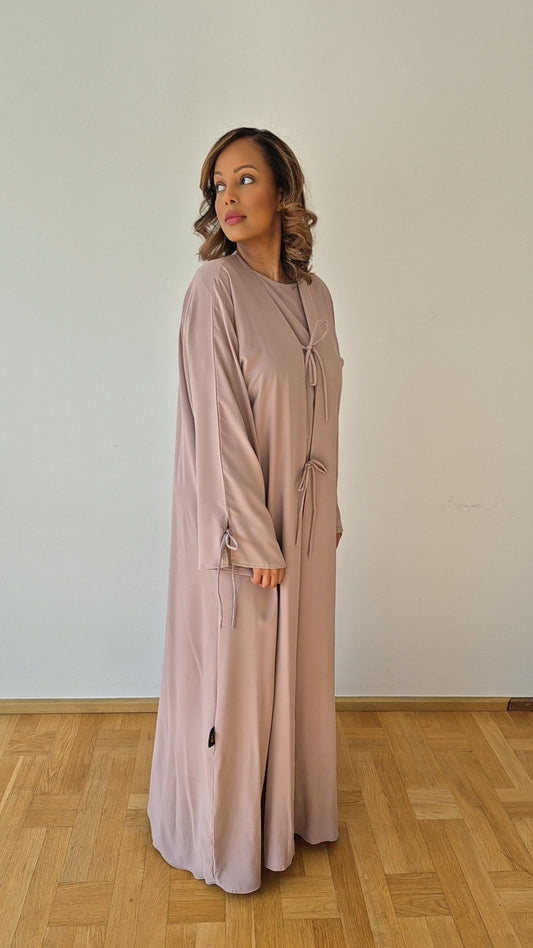 A woman stands on a wooden floor against a white wall, wearing the Bihi Boutique Shop Ayan Nude Nidah Dress (Size 60) with a chiffon hijab. She looks to the side with a neutral expression.