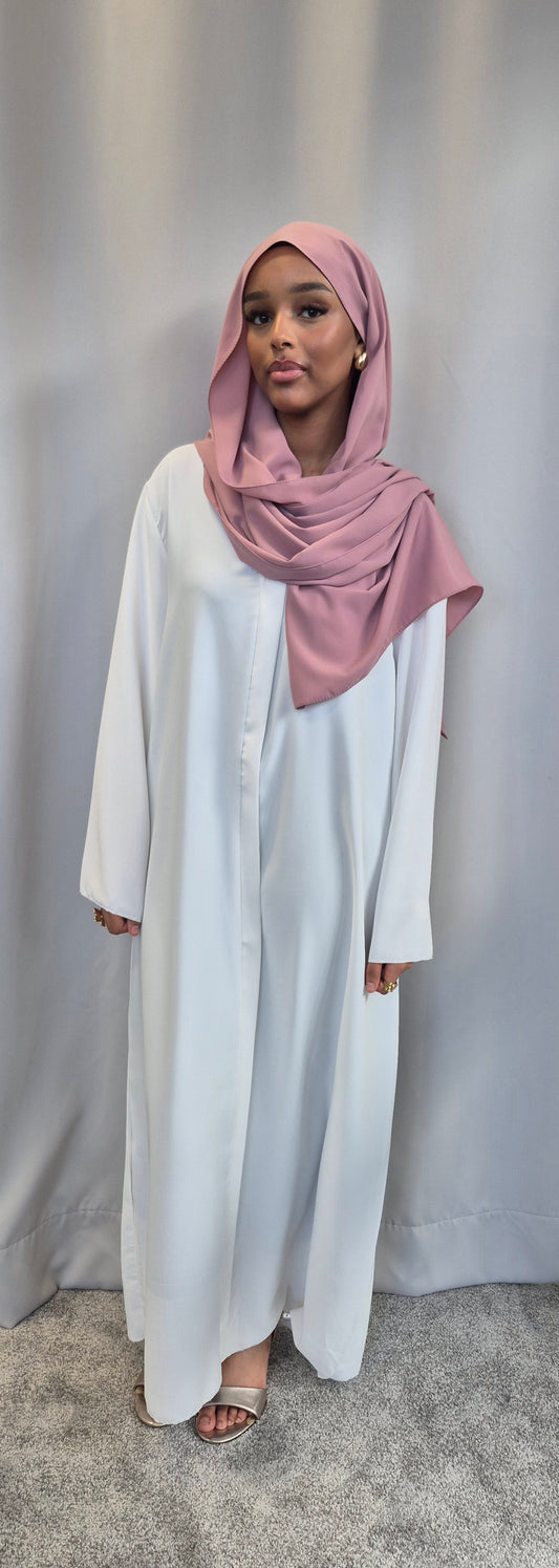 A woman stands on a grey carpet before a grey curtain, wearing the Bihi Boutique Islene Dress in white (one size fits 160-165 cm) and a pink hijab. She has a neutral expression and is wearing sandals.
