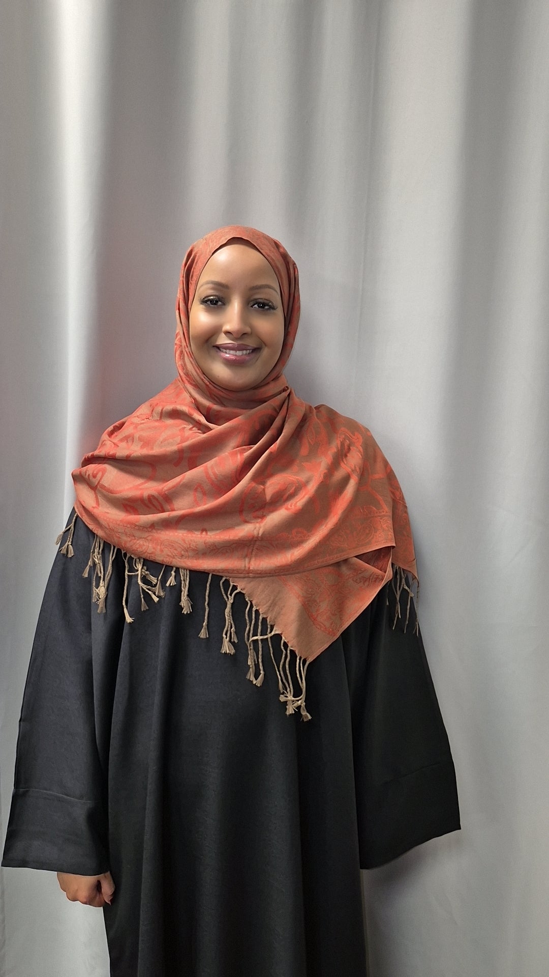 A woman in a burnt orange Luxurious Pashmina Hijab by Bihi Boutique and a long black dress smiles in front of a light gray curtain.