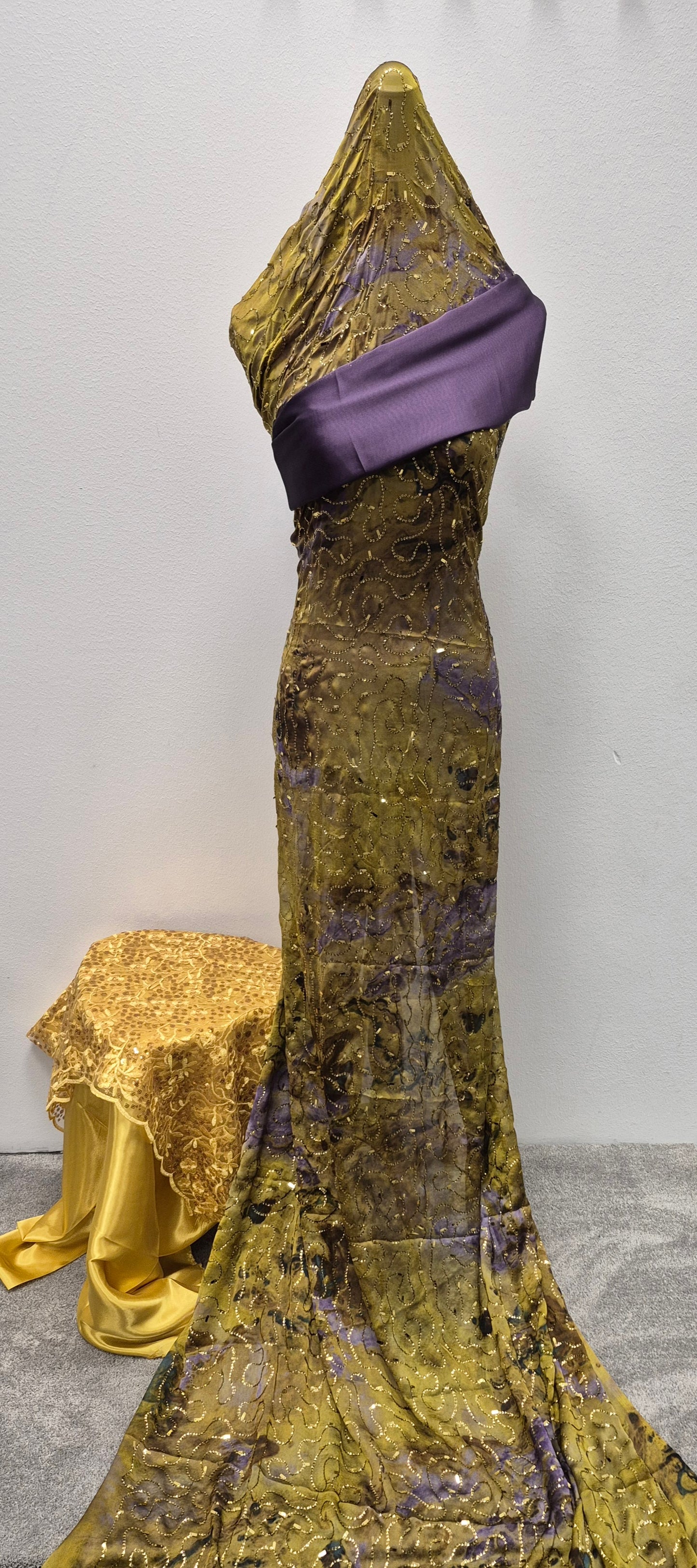 A mannequin showcases the Shop Fransawi Dirac Half Bac - Elegant Traditional Wear by Bihi Boutique in shimmering gold and purple, accented with a wide sash. Behind, a matching gold and yellow draped cloth decorates a low table.