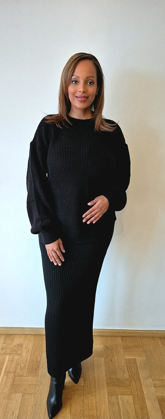 A woman smiles indoors against a white wall, wearing the Chic Black Sweater & Skirt Set by Bihi Boutique with black boots. One hand rests on her abdomen, and she stands on a wooden floor.