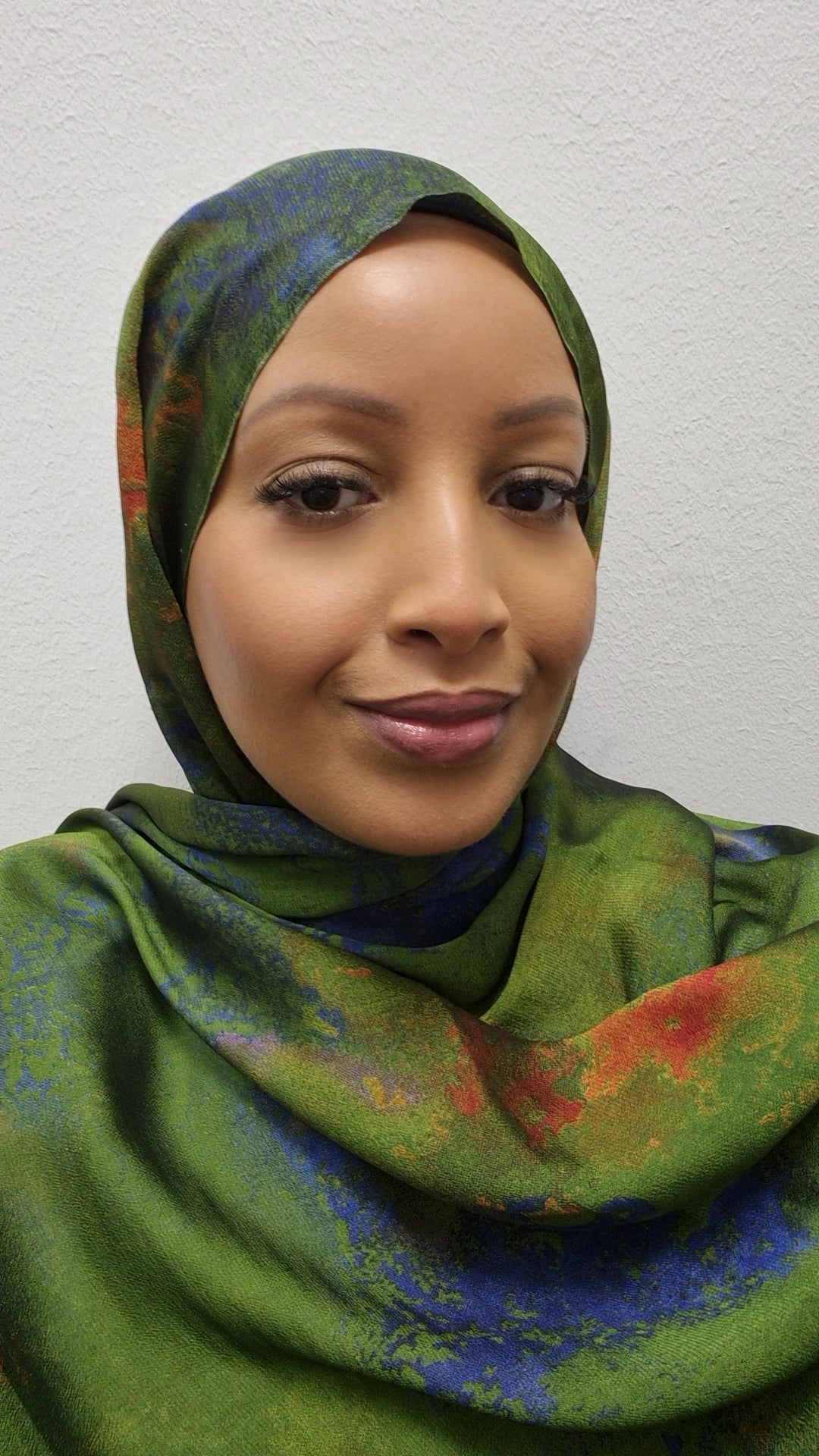 A woman models the Elegant Printed Chiffon Hijab from Bihi Boutique, featuring blue and orange patterns on green, as she smiles gently against a plain, light-colored wall.