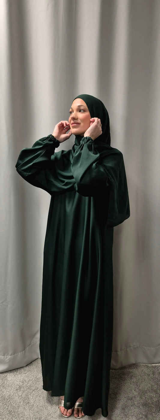 A woman stands indoors on a light carpet, wearing the dark green Shop Amina Abaya with Chiffon Hijab (Size 56) from Bihi Boutique, gently touching her headscarf. A light gray curtain is in the background.