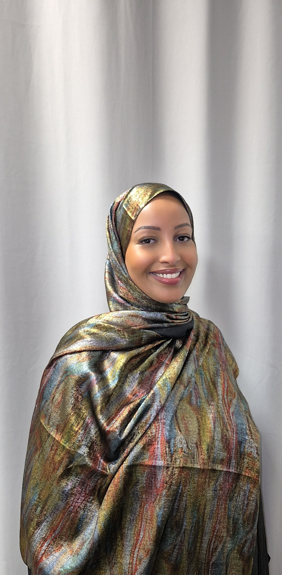 A woman smiles warmly at the camera, wearing Bihi Boutiques Elegant Satin Hijab—luxuriously soft and stylish—paired with matching attire, standing in front of a plain light curtain backdrop.