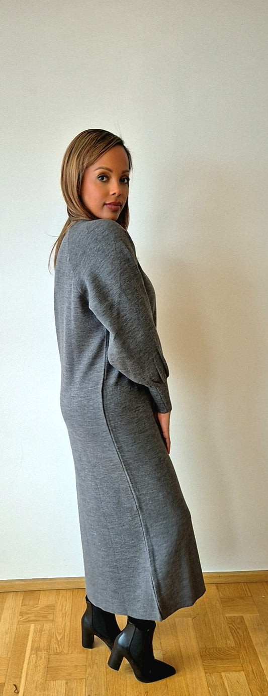 A woman poses indoors on a wooden floor, wearing the Bihi Boutique Shop Stylish Grey Sweater Dress from the Cozy Winter Collection, featuring puffed sleeves and paired with black heeled boots, against a plain light wall.