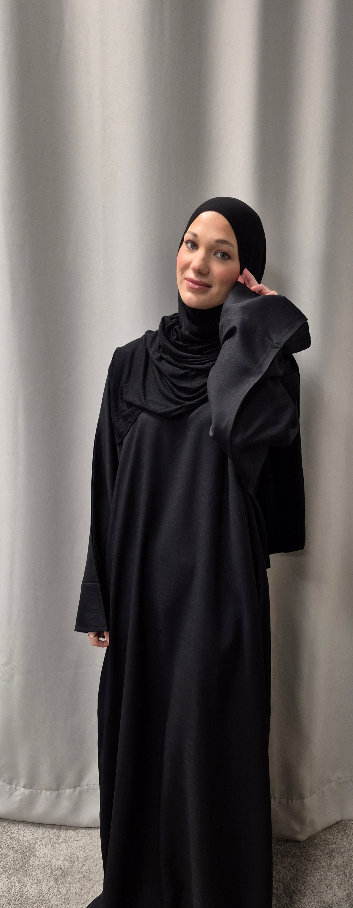A woman models the Elegant Linen Zamzam Black Abaya (Size 54) by Bihi Boutique, smiling softly with her hand on her face in front of a light gray curtain. Shop now!.