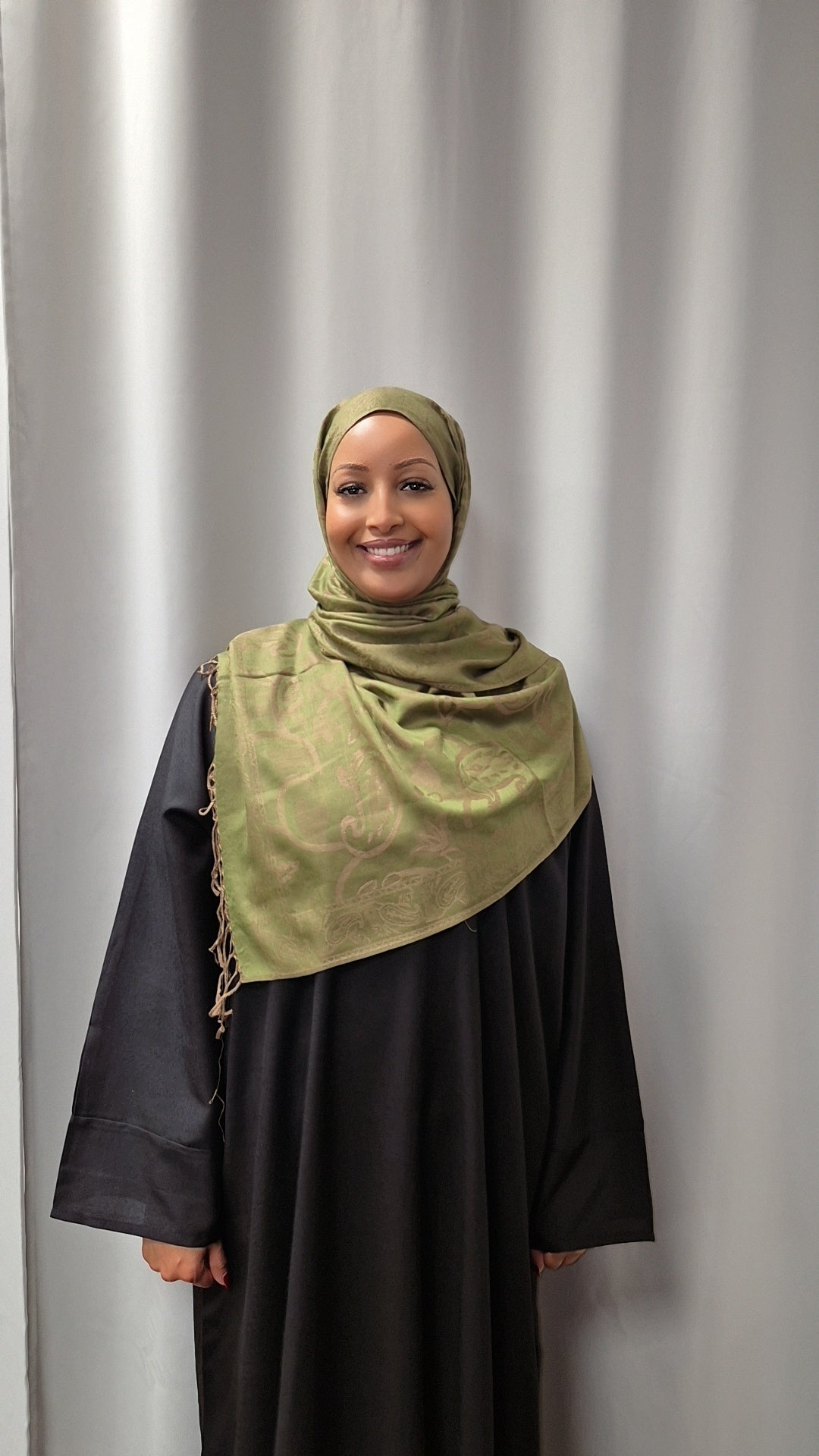 A woman smiles in front of a light gray curtain, wearing a black outfit paired with Bihi Boutiques Luxurious Pashmina Hijab in olive green—a soft, stylish, and versatile accessory.