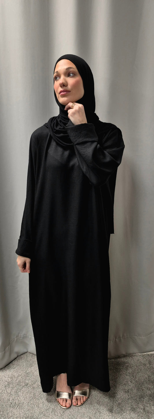 A person wearing the Chic Linen Abaya in Black with Pockets (Size M) from Bihi Boutique stands on a gray carpet in front of light curtains, looking thoughtfully to the side with one hand near their chin.