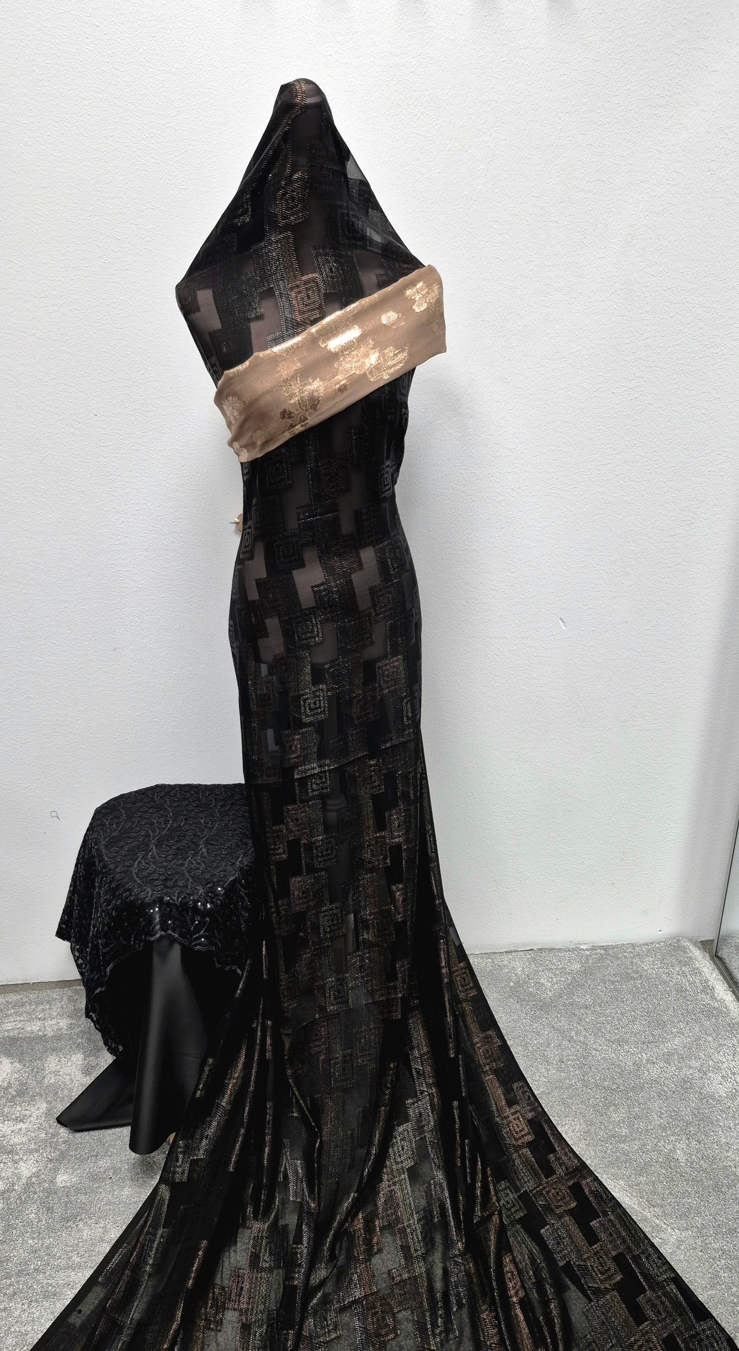 A mannequin showcases Bihi Boutique’s Shop Fransawi Dirac: an elegant, long black patterned gown with a geometric metallic design and a shiny taupe sash across the bodice. Display set against a plain white background and grey carpeting.