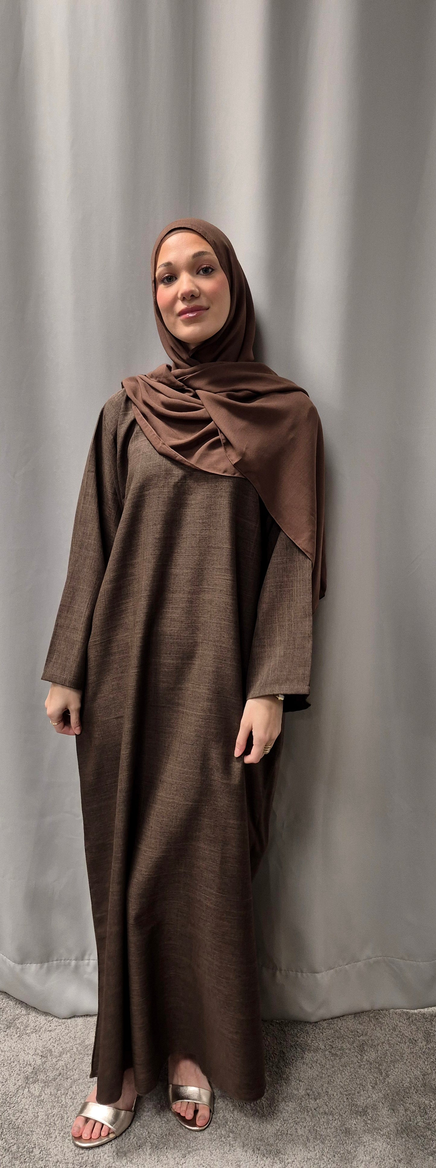 A woman smiles gently at the camera in front of a light gray curtain, wearing Bihi Boutique’s Stylish Linen Zamzam Brown Abaya with Chiffon Hijab (Size 52) and white sandals.