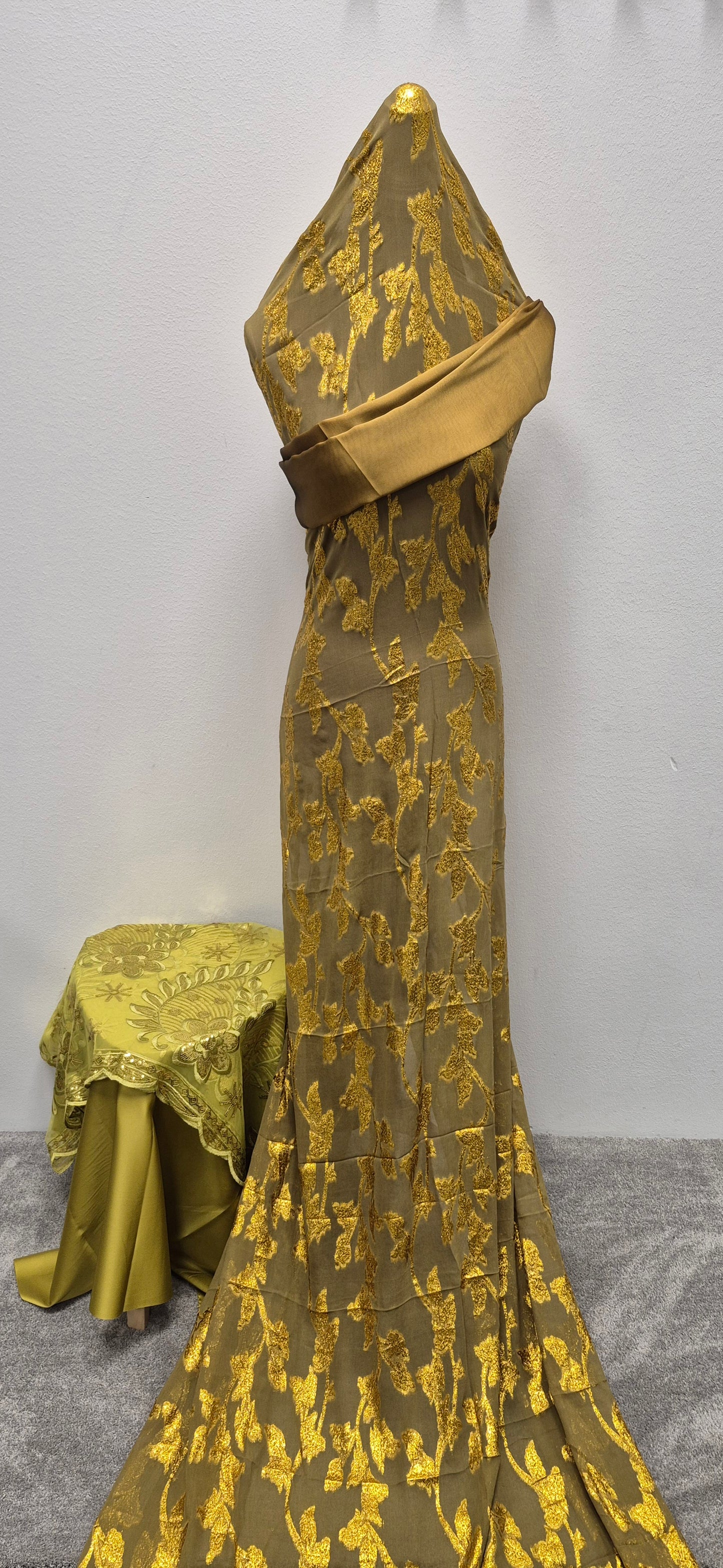 A mannequin showcases the Shop Fransawi Dirac Half Bac by Bihi Boutique, featuring elegant gold and olive green floral brocade, draped over the upper body and floor. A nearby small table is also covered in this stylish matching fabric.