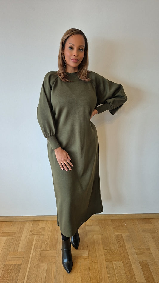 A woman stands indoors against a plain white wall, modeling the Olive Green Sweater Dress by Bihi Boutique—modest winter wear for women. The long-sleeve knit dress is paired with black ankle boots, and she smiles softly with one hand on her hip.