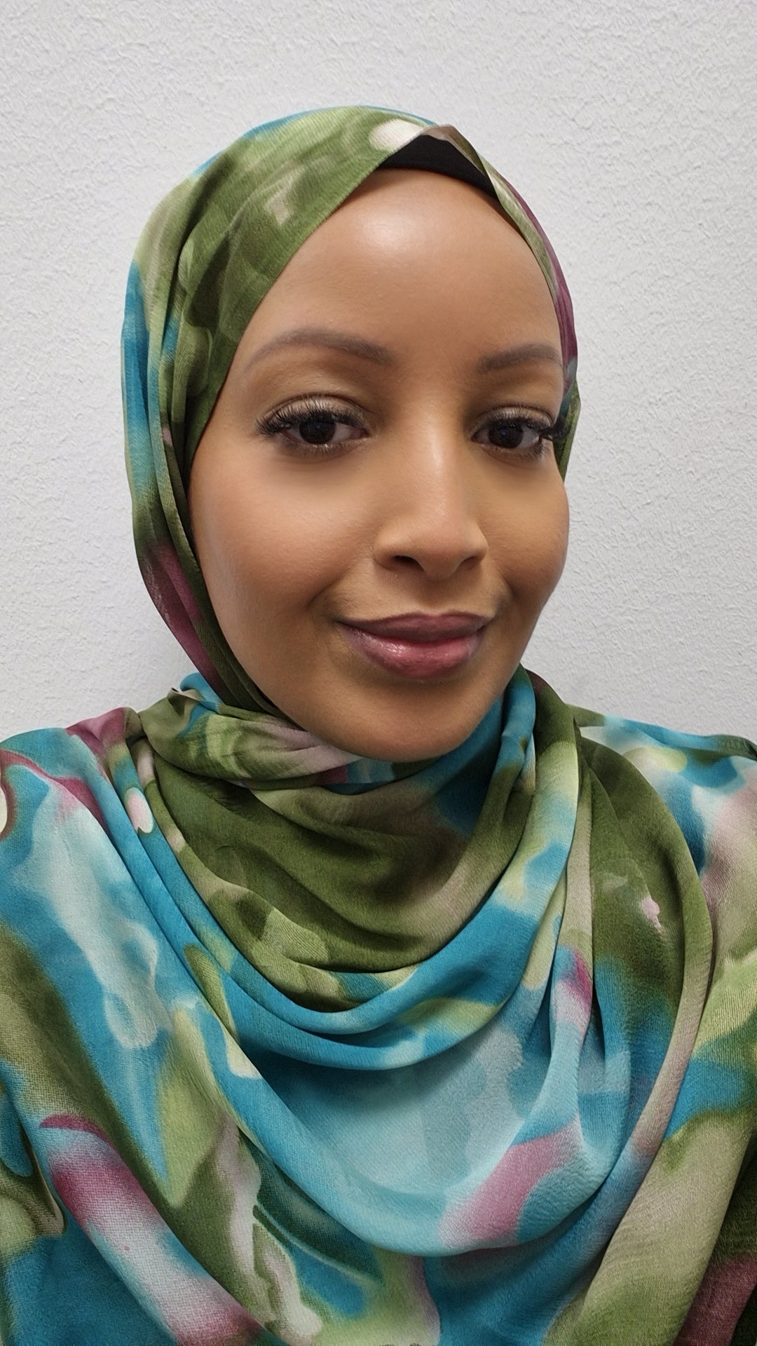 A woman in Bihi Boutiques Elegant Printed Chiffon Hijab—lightweight and stylish with vibrant florals—and natural makeup, smiles softly at the camera against a plain, light-colored wall.
