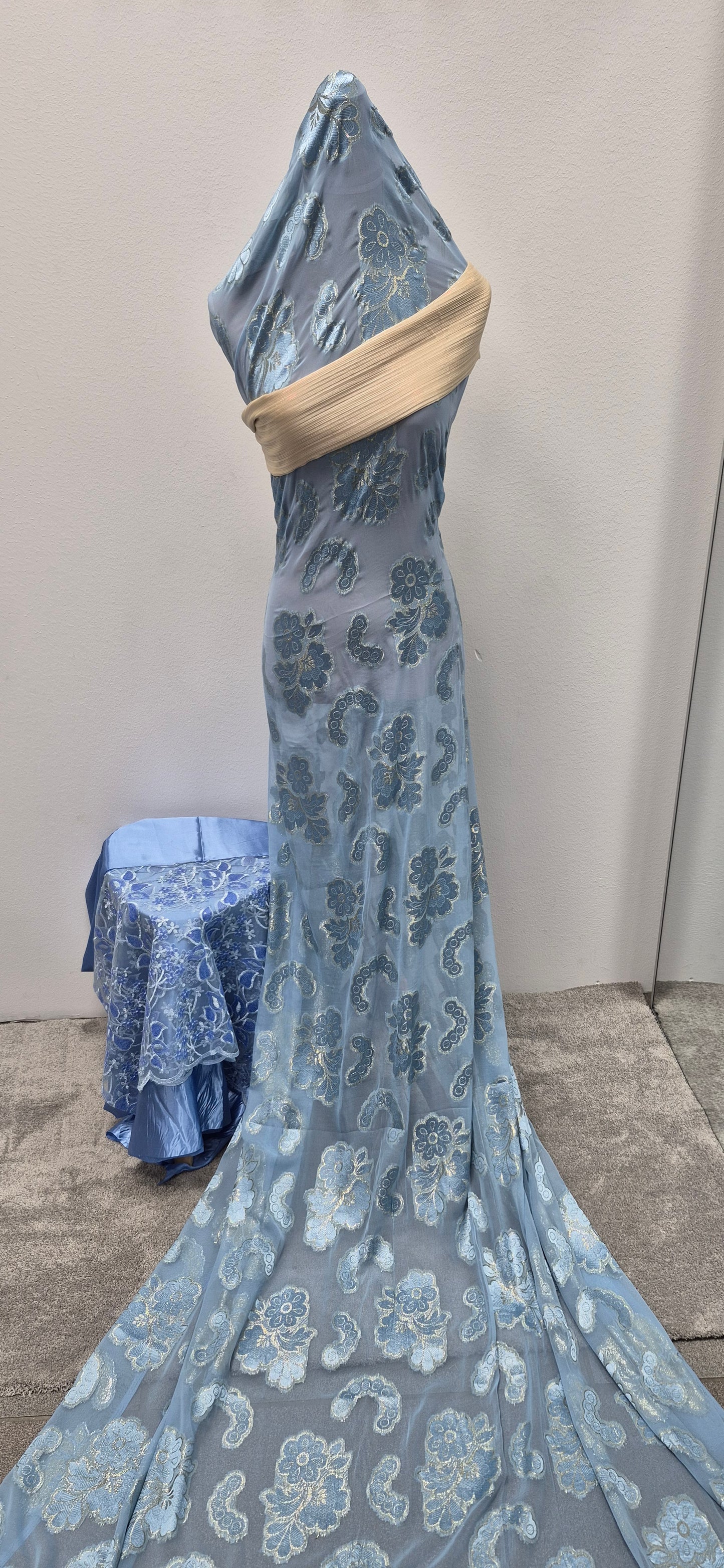 The Fransawi Dirac Bacweyne by Bihi Boutique, an elegant blue sheer Somali dress with embroidered floral patterns and a beige sash, is displayed on a mannequin beside a table draped in matching fabric, set against a plain background and carpeted floor.