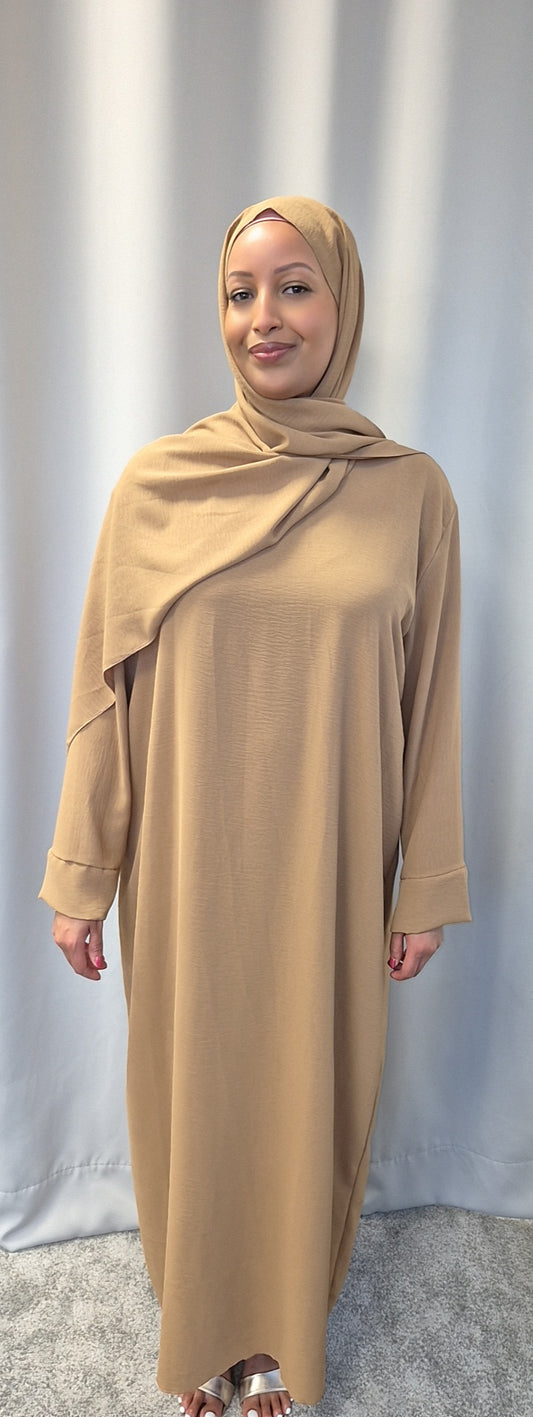 A woman stands in front of a light gray curtain, smiling gently as she wears the Elegant Black One Size Prayer Clothing from Bihi Boutique, with her hands relaxed by her sides and light sandals on a gray carpet.