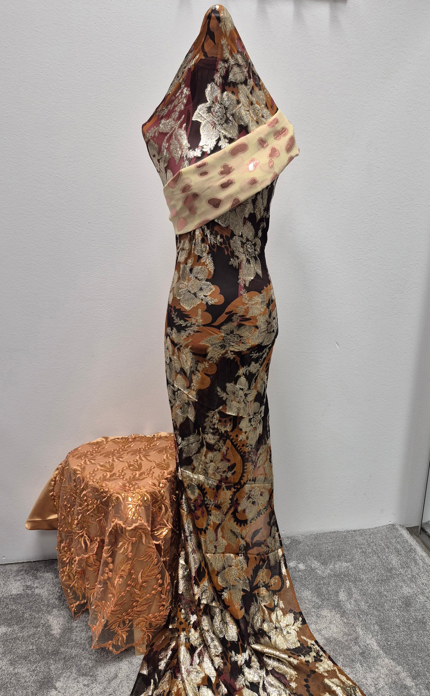 A mannequin showcases the Shop Stylish Fransawi Dirac (Bacweyne) by Bihi Boutique, a long fitted gown with metallic gold, bronze, and silver floral patterns and a sheer shawl. Nearby, a small table is draped in matching ornate fabric.