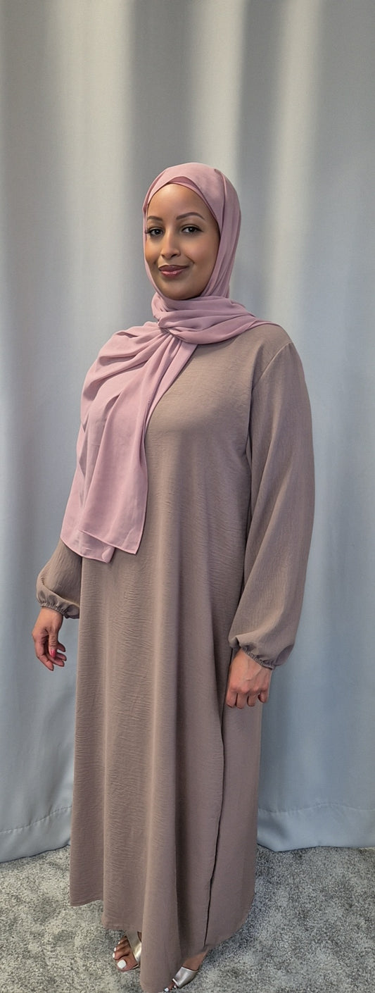 A woman in the Shop Elegant Nude Slip Dress from Bihi Boutique and a light pink hijab smiles at the camera, standing before a light gray curtain.