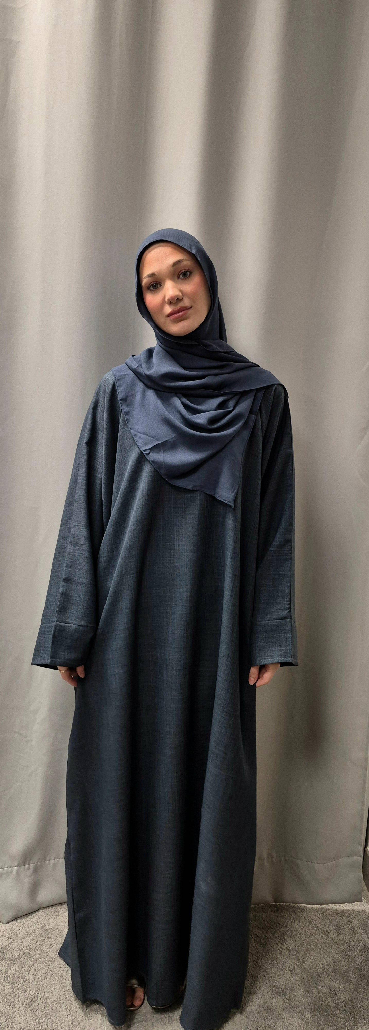 A woman models the Bihi Boutique Linnen Zamzam Blue Size 56 abaya and matching hijab—an elegant, lightweight fashion piece—standing before a gray curtain and gazing neutrally at the camera.