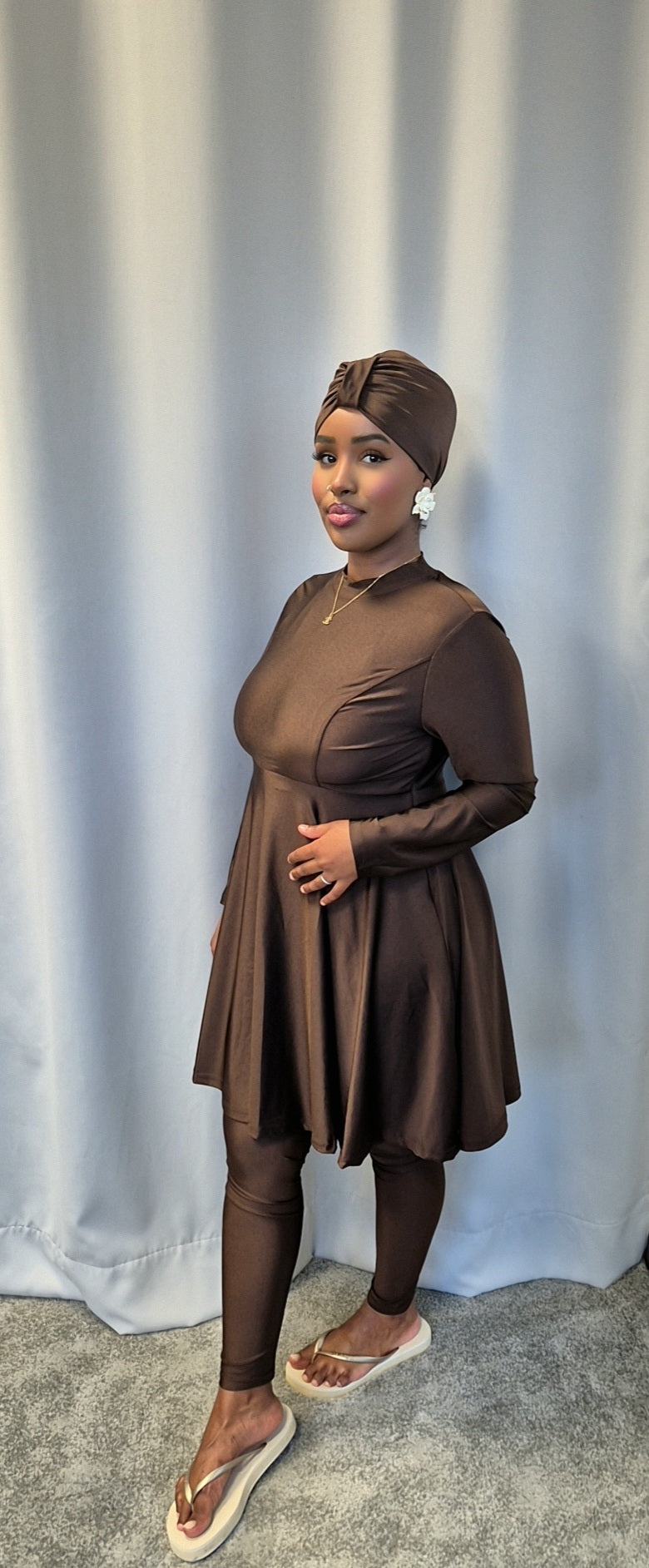 A woman models the Shop Stylish Black Burkini from Bihi Boutique, standing on a gray carpet before a light gray curtain and smiling softly.