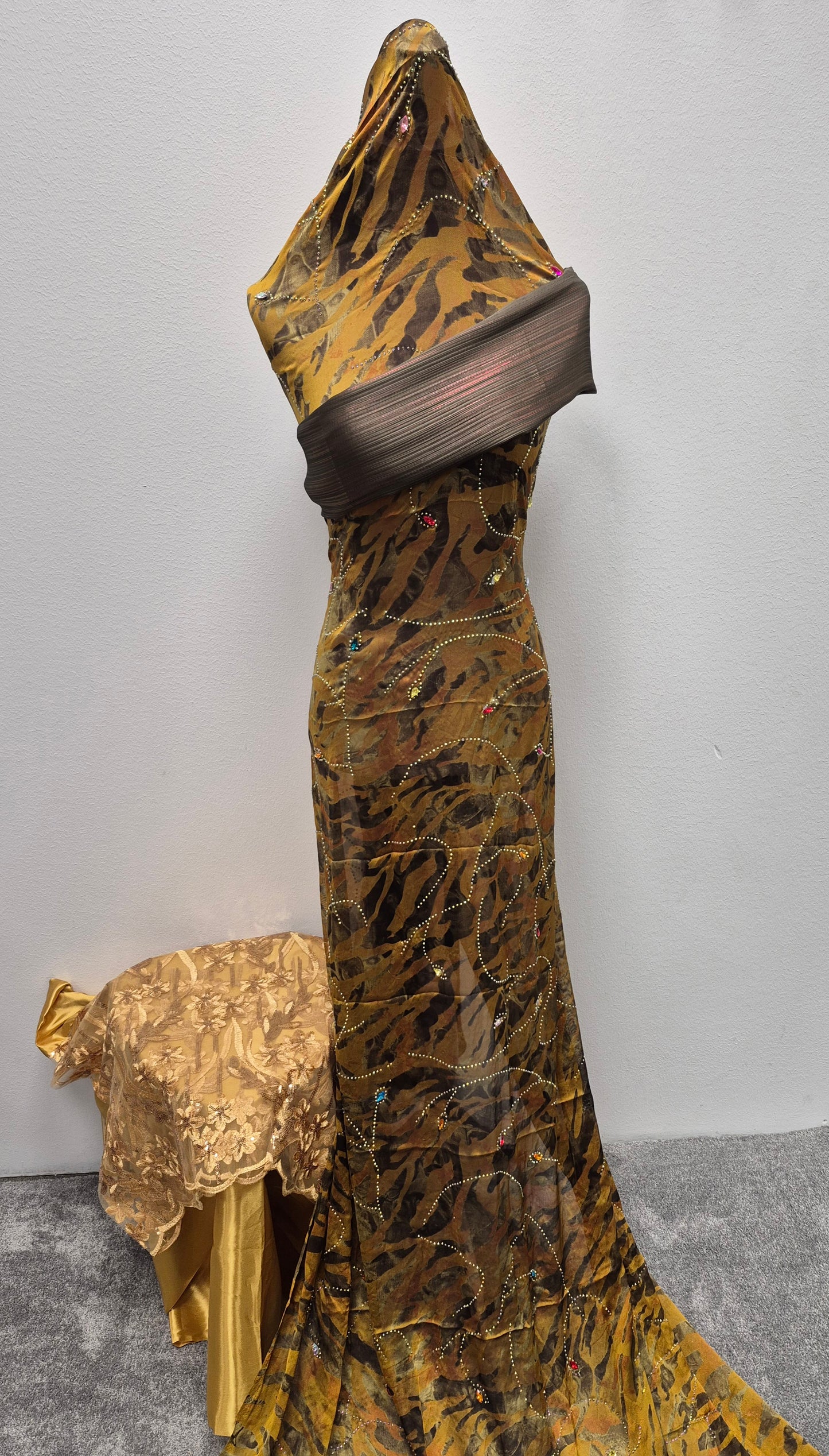 A mannequin models the Shop Fransawi Dirac Half Bac - One Size Elegance by Bihi Boutique, a floor-length, sheer brown and gold tiger-striped dress with sequins and a mesh bodice. A nearby table features a gold-patterned cloth, set against a white background.