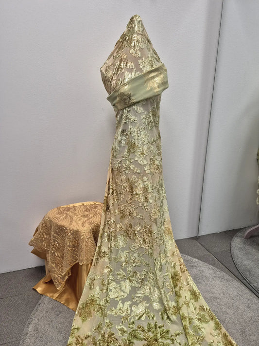 The Magbal Dirac Half Bac Dress by Bihi Boutique, a pale gold floor-length gown with intricate floral embroidery, is showcased on a dress form beside a table draped in matching fabric, set against a neutral wall and gray carpet.