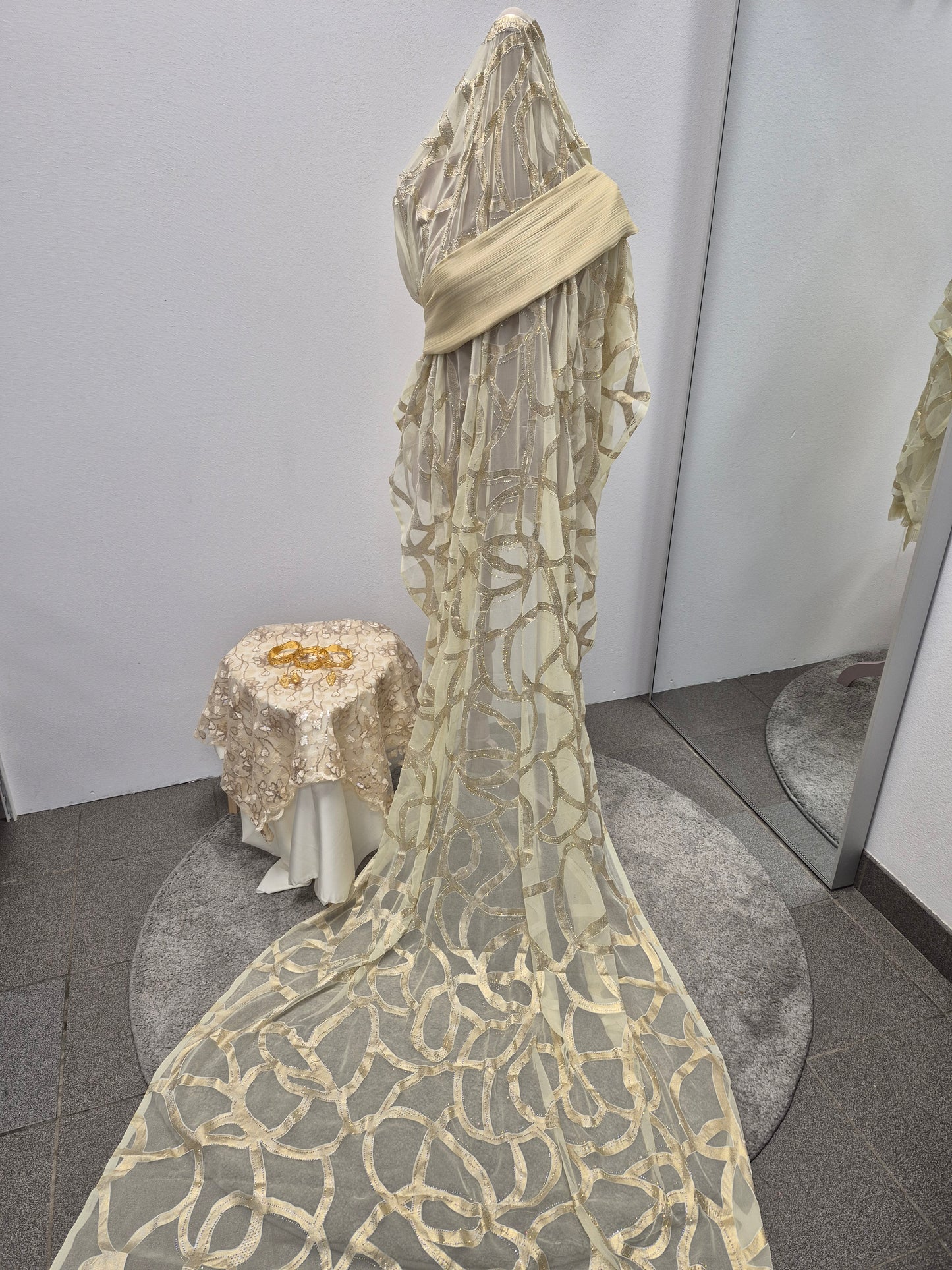 A mannequin displays the Shop Nikah Fransawi Dirac - Elegant Gyllene Half Bac Dress by Bihi Boutique, crafted from sheer cream fabric with circular patterns and a long train. Nearby, gold jewelry sits on a matching table beside mirrored walls.