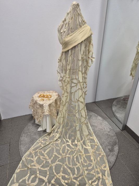 A mannequin displays the Shop Nikah Fransawi Dirac - Elegant Gyllene Half Bac Dress by Bihi Boutique, crafted from sheer cream fabric with circular patterns and a long train. Nearby, gold jewelry sits on a matching table beside mirrored walls.