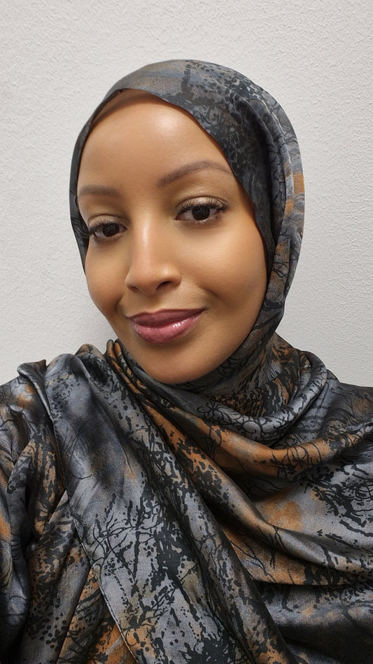 A woman smiles gently at the camera, wearing the Elegant Chiffon Hijab in gray and brown from Bihi Boutique—a soft, stylish headscarf for all occasions—while standing against a plain white wall.