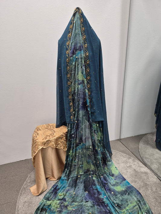 A mannequin displays the Bihi Boutique Fransawi Dirac Half Bac Dress for Women, featuring deep blue and green ornate fabric with gold trim and sequins, seated on a beige patterned stool as part of the dress trails elegantly onto the floor.