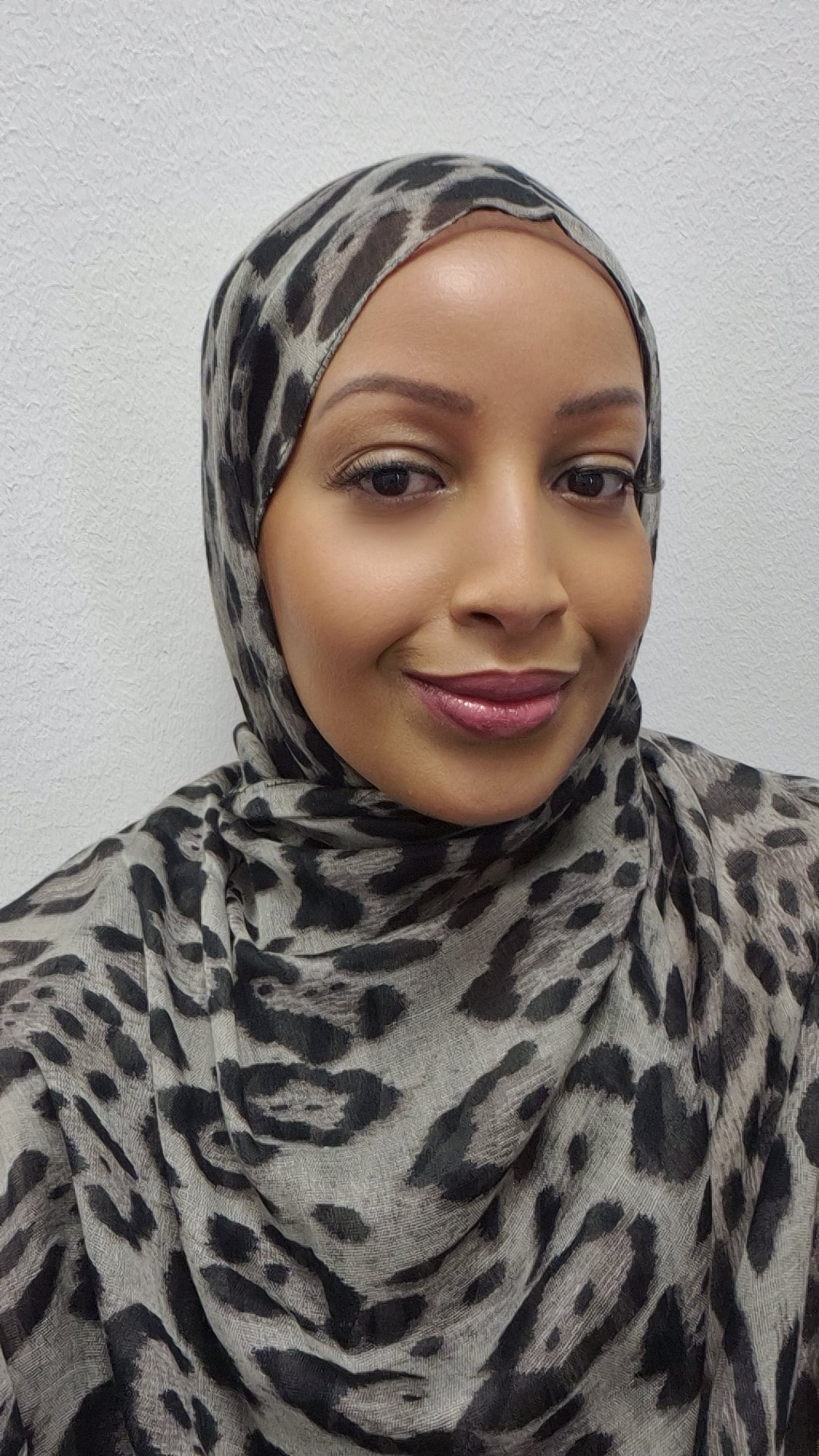 A woman wearing the Stylish Cheetah Hijab by Bihi Boutique smiles softly at the camera, posed against a plain white wall.