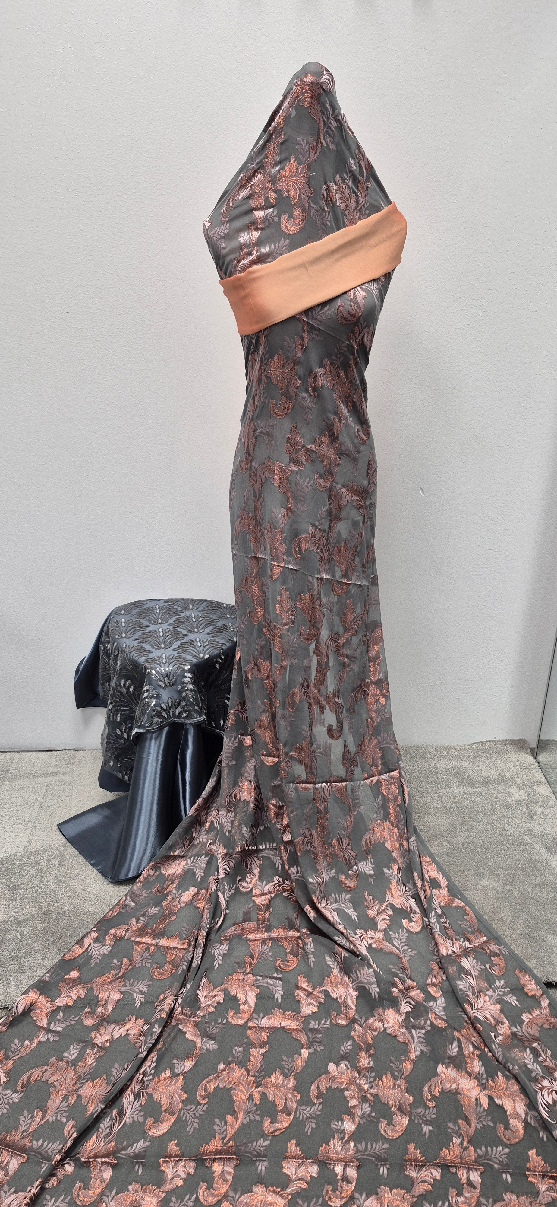 A headless mannequin showcases the Shop Elegant Fransawi Dirac Bacweyne by Bihi Boutique, featuring gray and pink patterns with a peach wrap. A dark, shiny fabric is displayed on a stand beside it, all set against a plain wall.