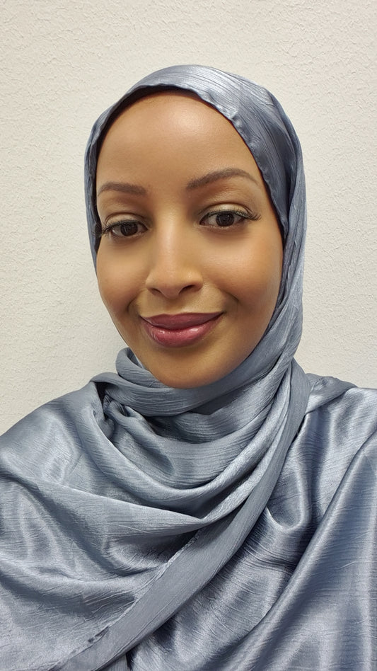 A woman wearing the Elegant Satin Hijab by Bihi Boutique in shiny light blue smiles gently at the camera, set against a plain white textured wall.