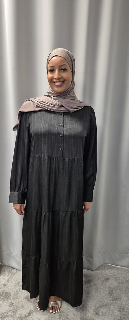 A woman in the Celeste Noir & Ivory Satin Dress by Bihi Boutique stands smiling on a light carpet in front of gray curtains, wearing a taupe hijab and open-toed sandals.