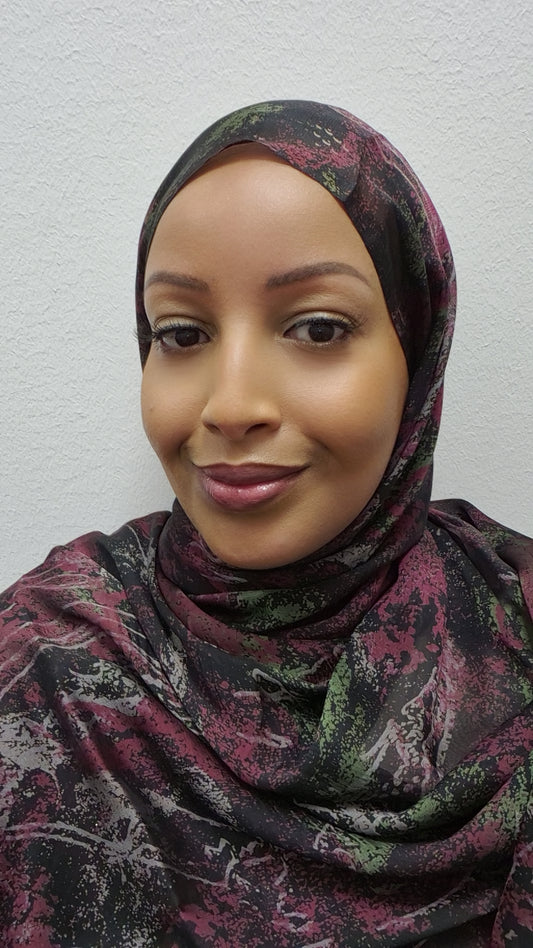 A woman wears the Elegant Chiffon Hijab by Bihi Boutique, featuring maroon, green, and black patterns. She smiles softly at the camera against a plain white wall background.