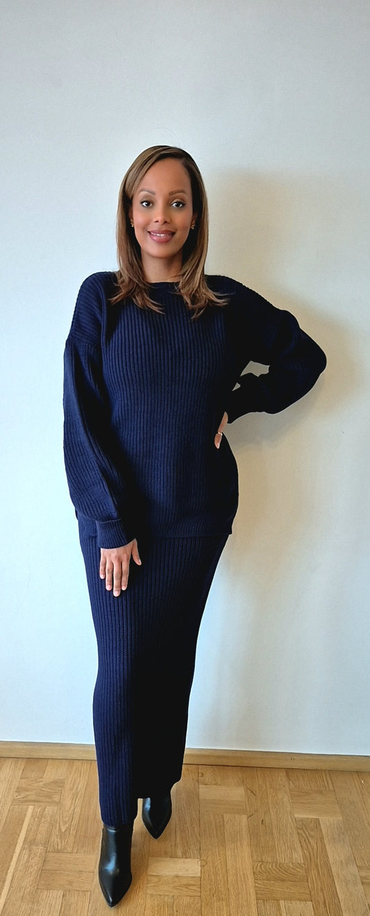 A woman stands indoors against a white wall, wearing the Navy Sweater & Skirt Set—a modest knit winter outfit from Bihi Boutique—with black ankle boots. She smiles and rests one hand on her hip.