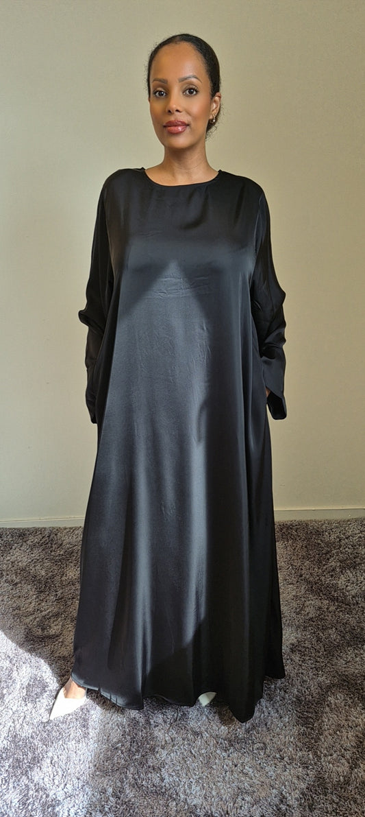 A person stands indoors on a carpet wearing the Elegant Satin Abaya Noireen by Bihi Boutique—a long, loose-fitting black evening dress with pockets and long sleeves. Light from the left highlights the outfit against a plain light wall.