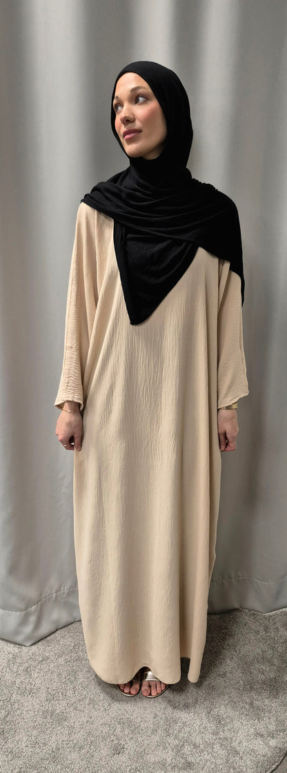 A woman wearing the Shop Gyllene Dress Size 60 from Bihi Boutique stands barefoot on a gray carpet before a light gray curtain, her black hijab complementing the elegant fashion as she gazes gently to her left.