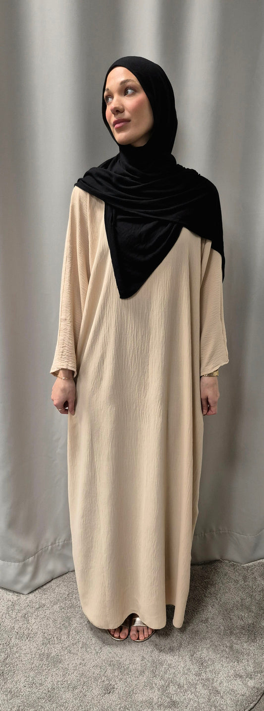 A woman wearing the Shop Gyllene Dress Size 60 from Bihi Boutique stands barefoot on a gray carpet before a light gray curtain, her black hijab complementing the elegant fashion as she gazes gently to her left.