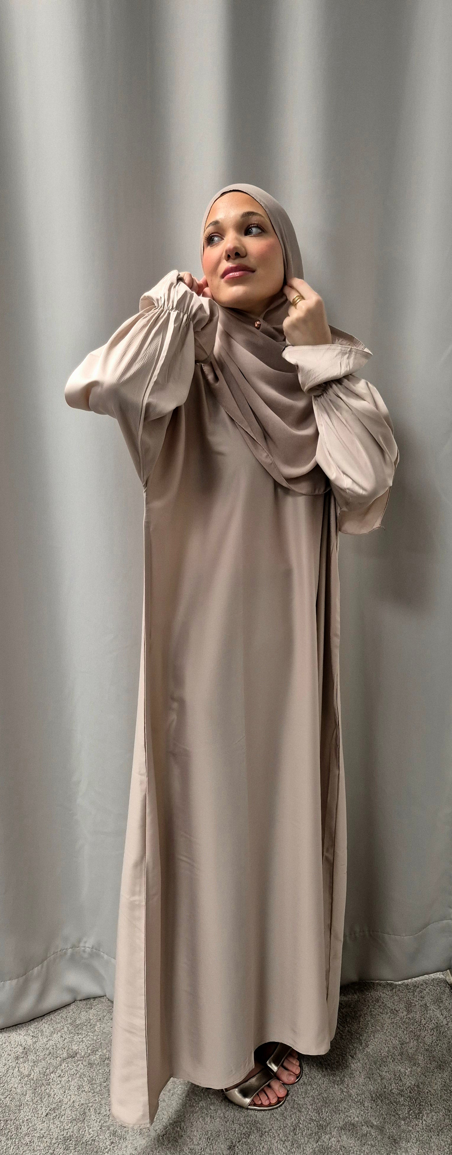 A woman wearing the Stylish Marwa Abaya Size 54 by Bihi Boutique with a matching hijab stands in front of gray curtains, looking upward as she adjusts her hijab, her sandals visible on a gray carpet.