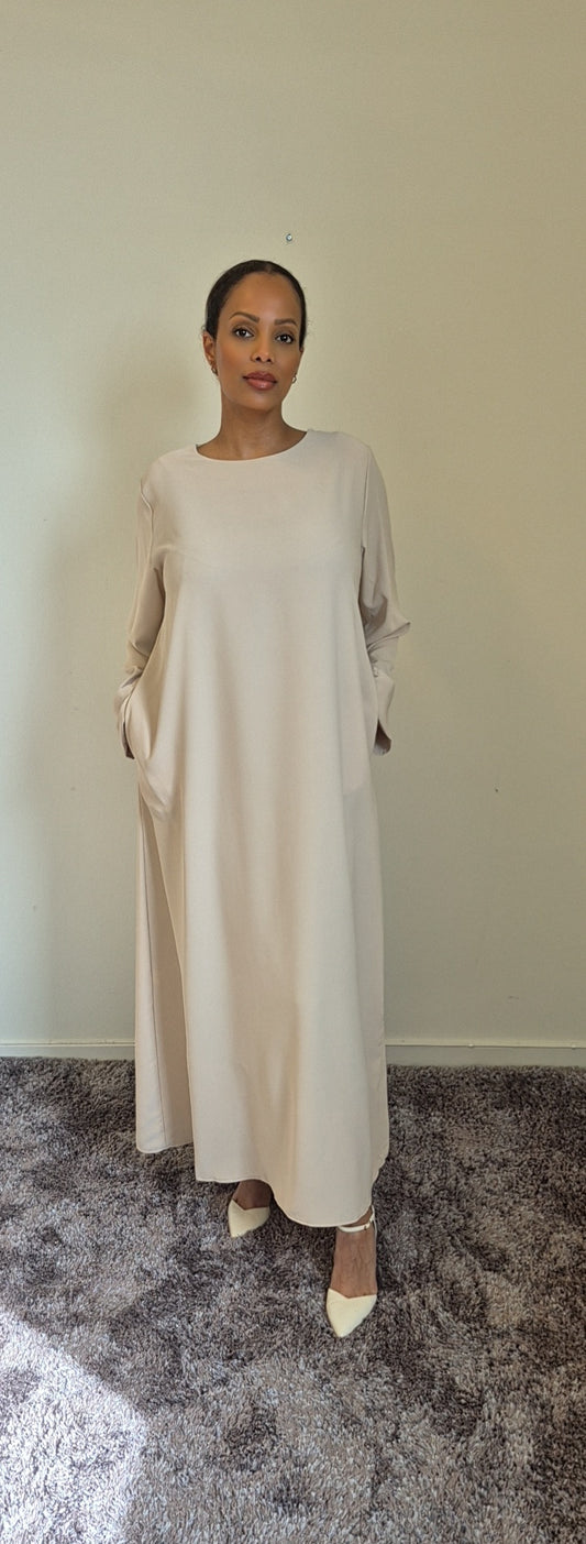 A woman stands indoors on a carpet, wearing the Elegant Black Hawra Abaya with pockets from Bihi Boutique. The long, loose abaya features long sleeves and combines style with versatility against a simple light background.
