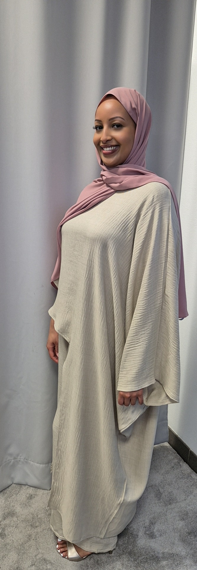A woman wearing the Shop Luxurious 2-Piece Linen Set from Bihi Boutique smiles in front of gray curtains, paired with a pink hijab and white heeled sandals, her hand relaxed at her side.