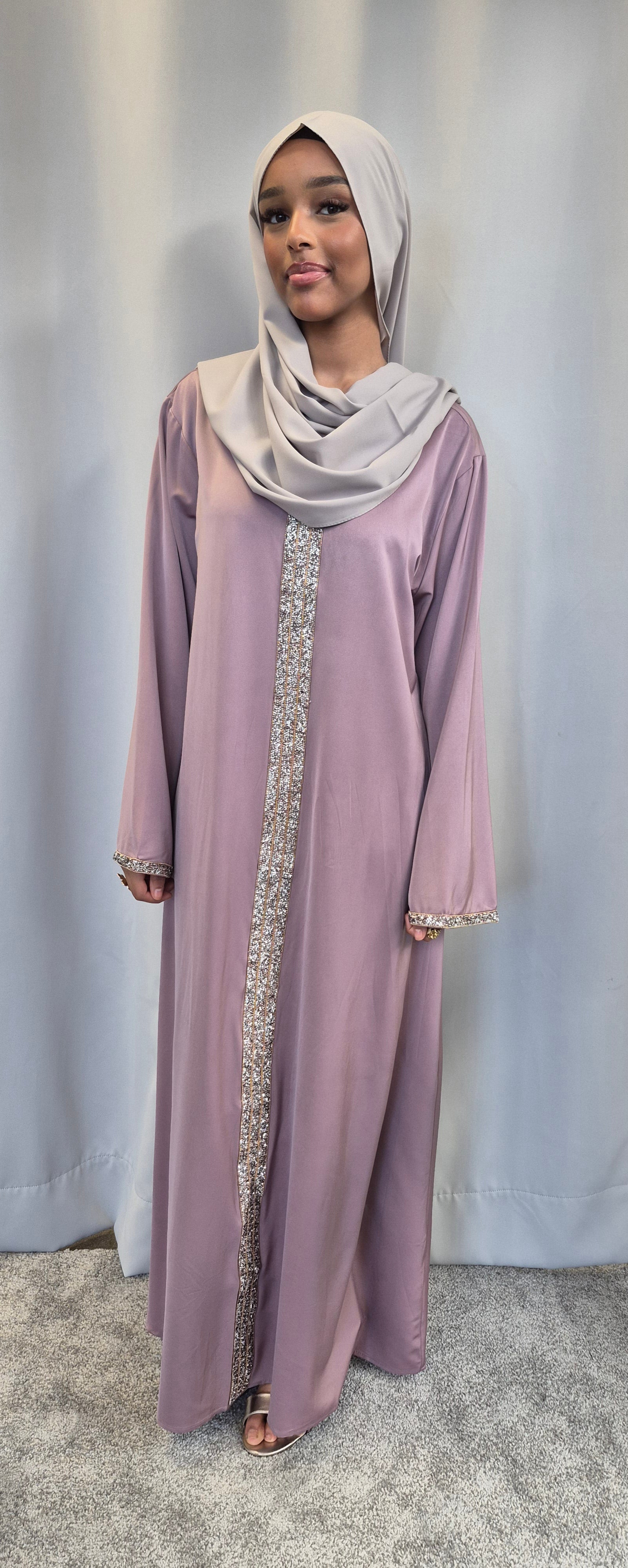 A woman in the Aurelia Ivory & Rose Dress by Bihi Boutique, an elegant S/M fashion essential, stands against a light grey backdrop, wearing a light grey hijab and looking at the camera with a gentle expression.
