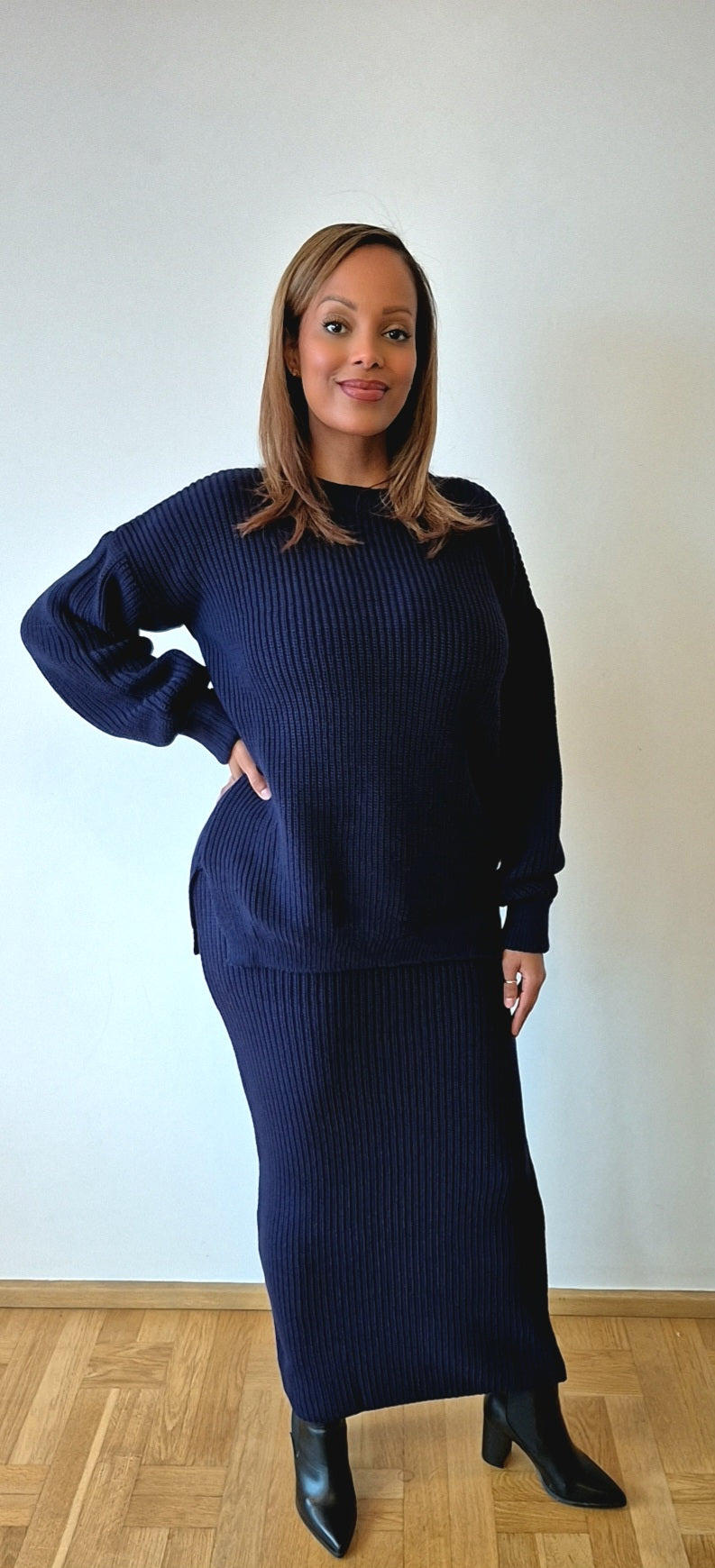 A woman stands indoors against a light wall, wearing the Navy Sweater & Skirt Set from Bihi Boutique. She has shoulder-length hair, black ankle boots, and looks at the camera with a slight smile.