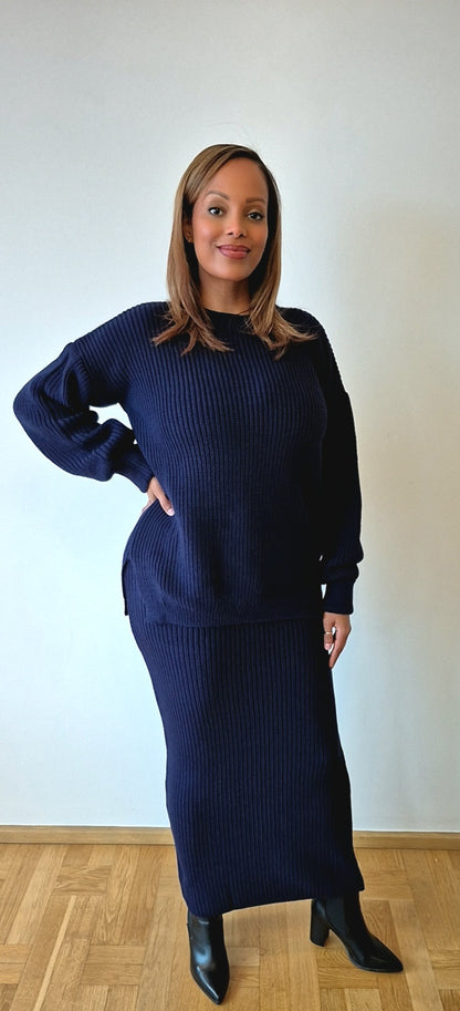 A woman stands indoors against a light wall, wearing the Navy Sweater & Skirt Set from Bihi Boutique. She has shoulder-length hair, black ankle boots, and looks at the camera with a slight smile.