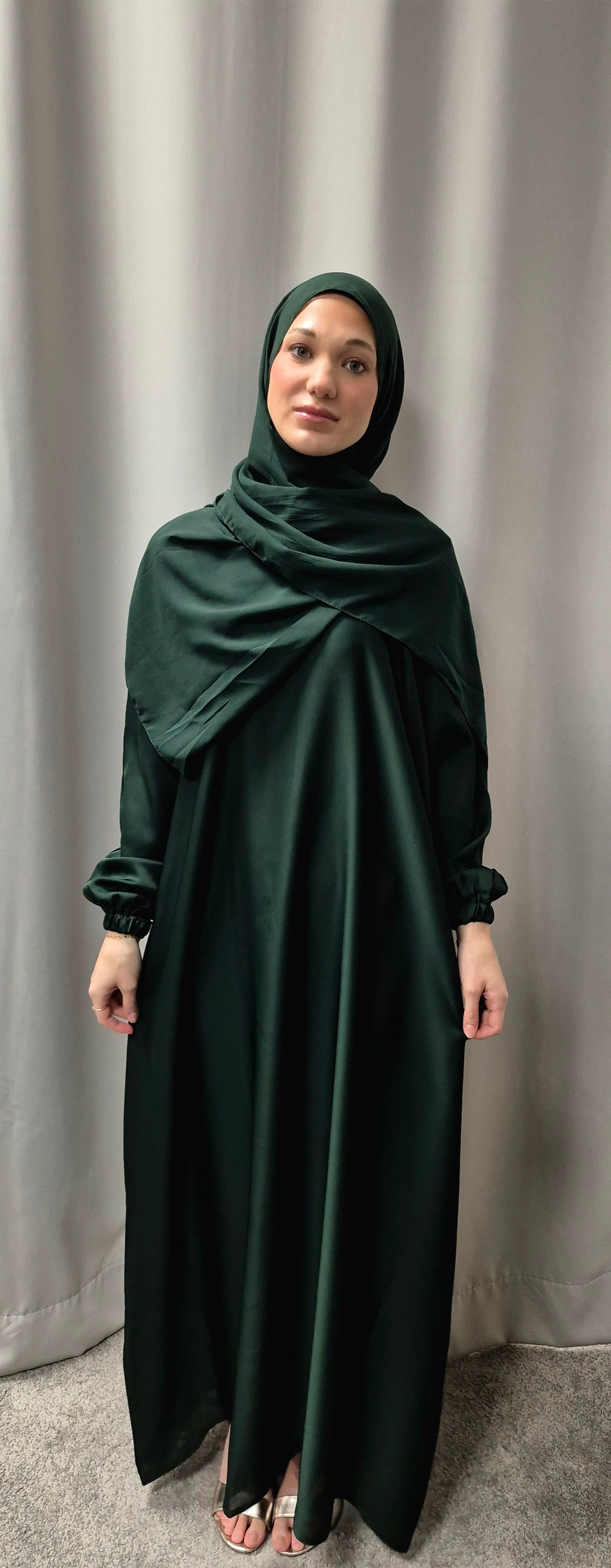 A woman stands before a light gray curtain, wearing the Shop Amina Abaya with Chiffon Hijab in Size 56 by Bihi Boutique. She faces forward with a neutral expression on a carpeted floor.